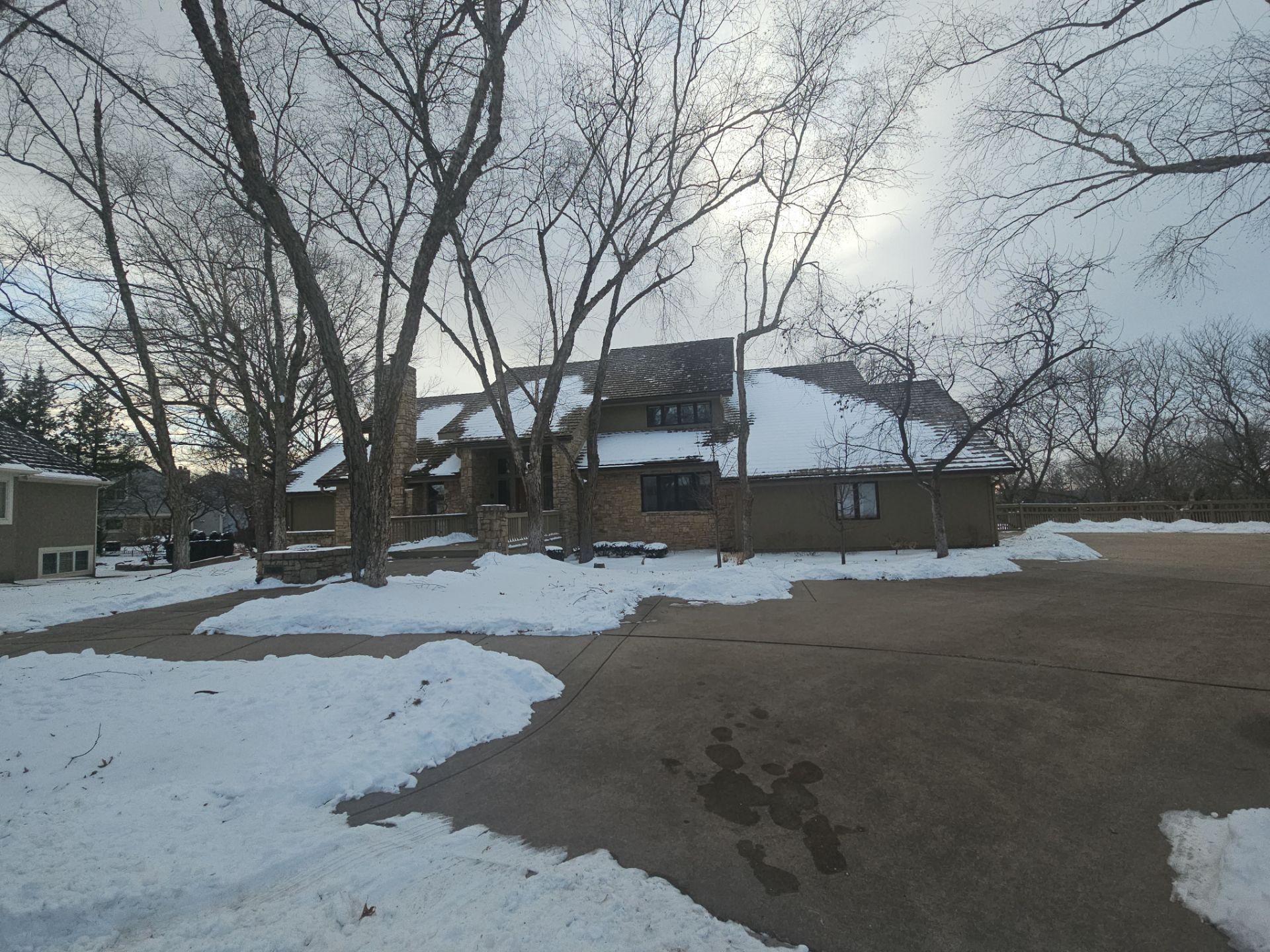 House with snow-covered roof and ground, trees with bare branches, driveway. Cloudy sky.