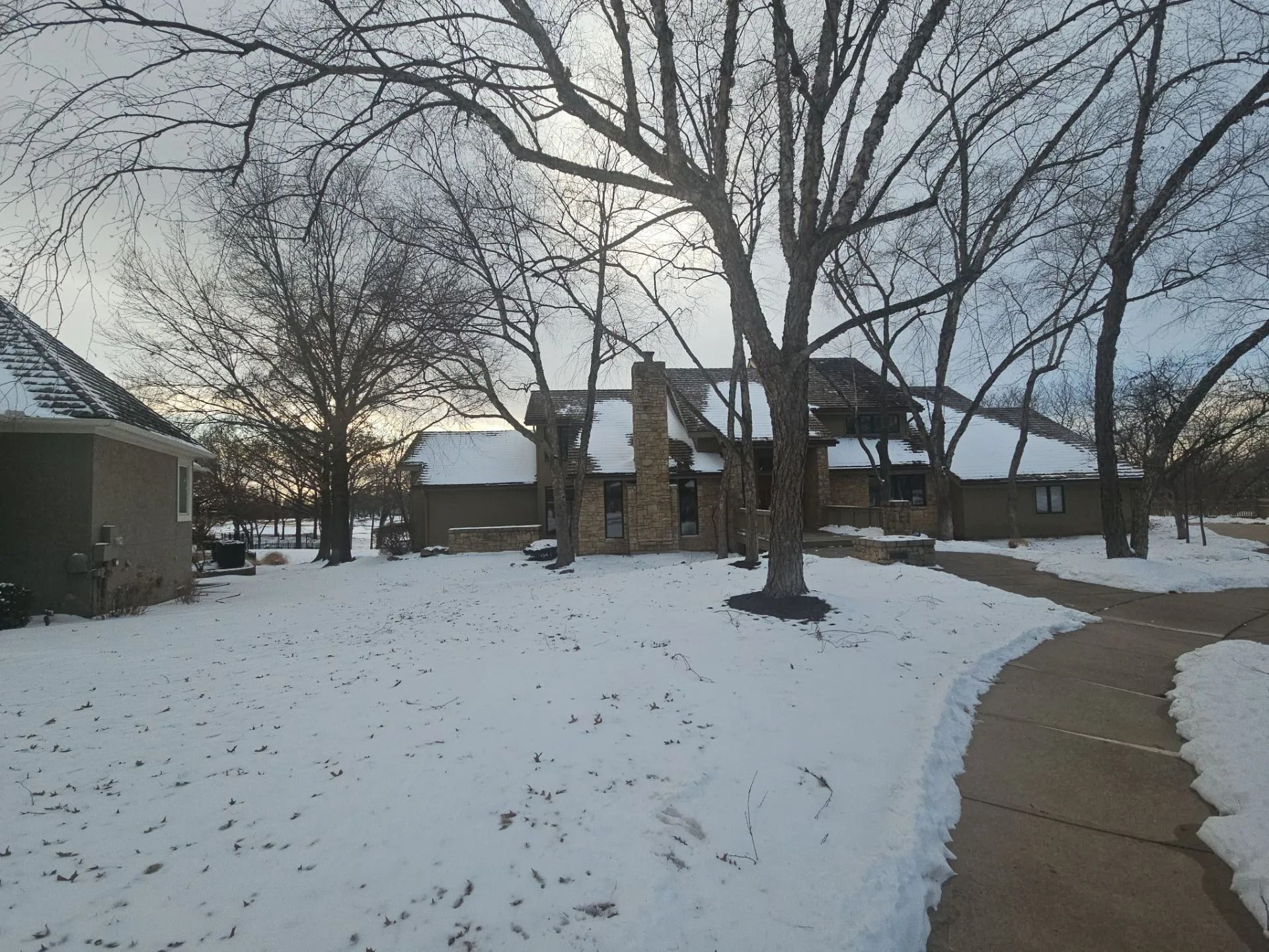 Snow-covered yard with a house and trees. A sidewalk is partially visible, leading towards the home. Cloudy sky.