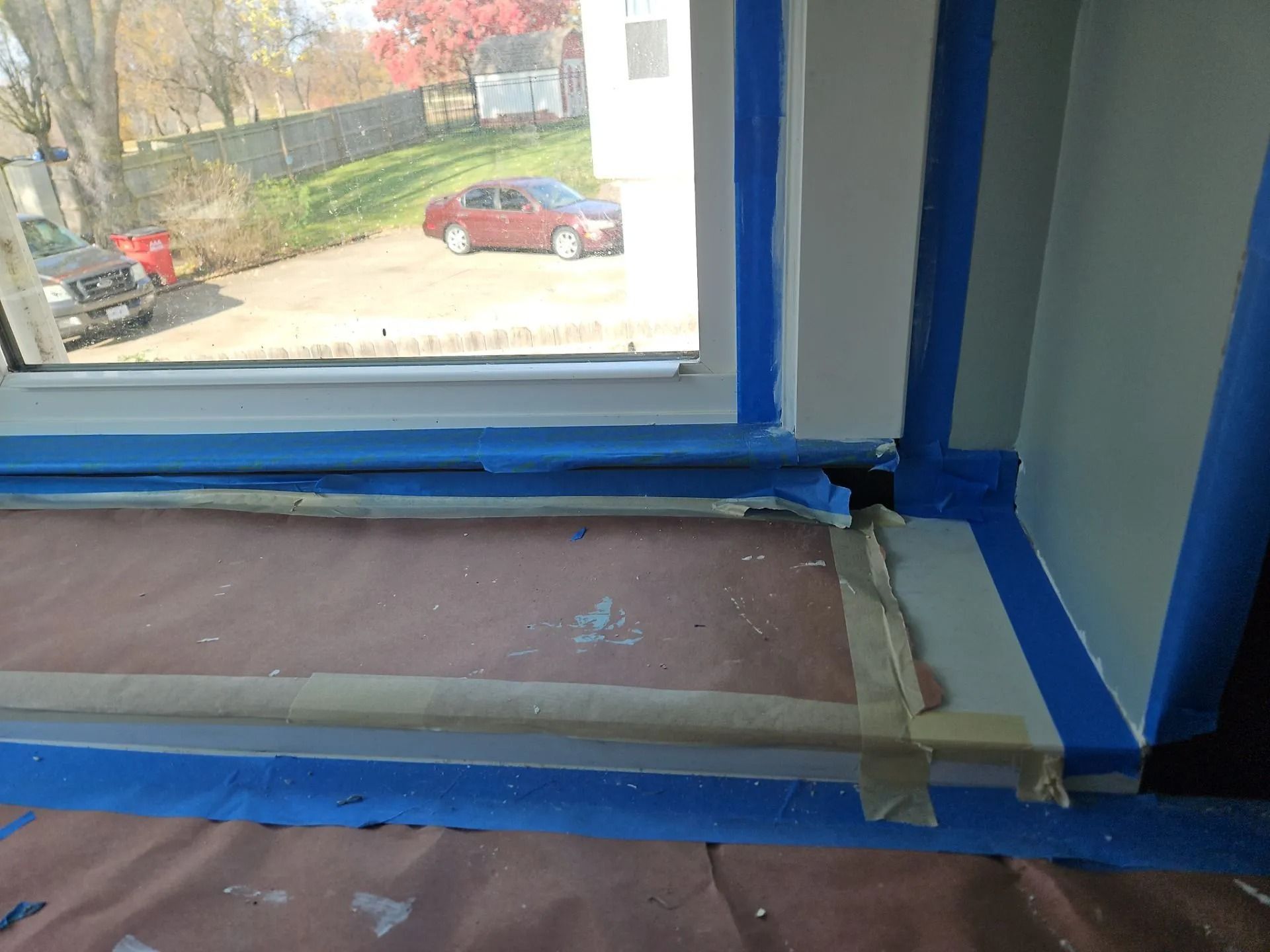 Window frame prepped for painting with blue tape; tan sill, vehicles outside.