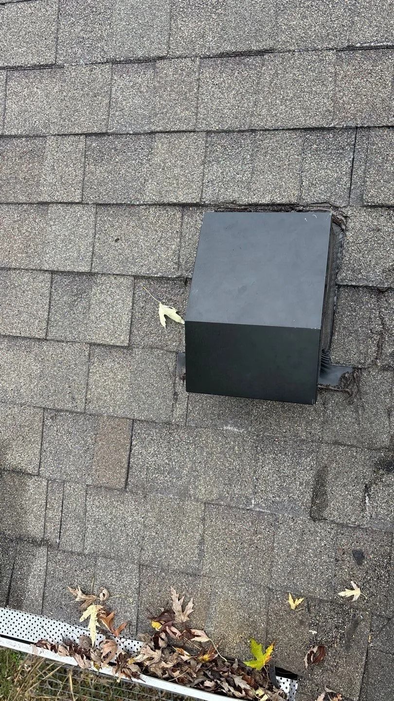 Black vent pipe on gray shingled roof, next to a gutter with leaves.