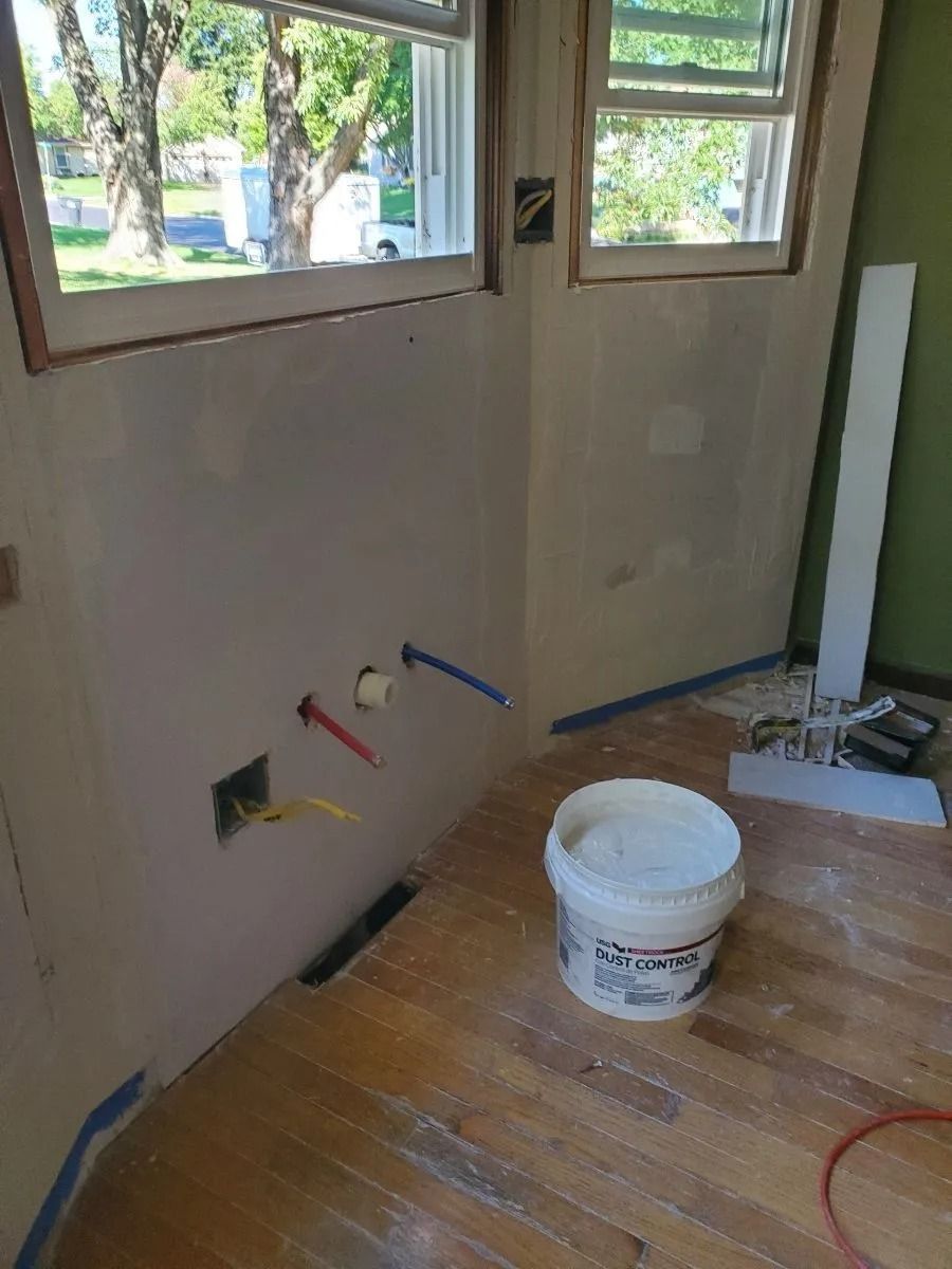 Renovated kitchen corner with exposed pipes, windows, and a bucket of joint compound.
