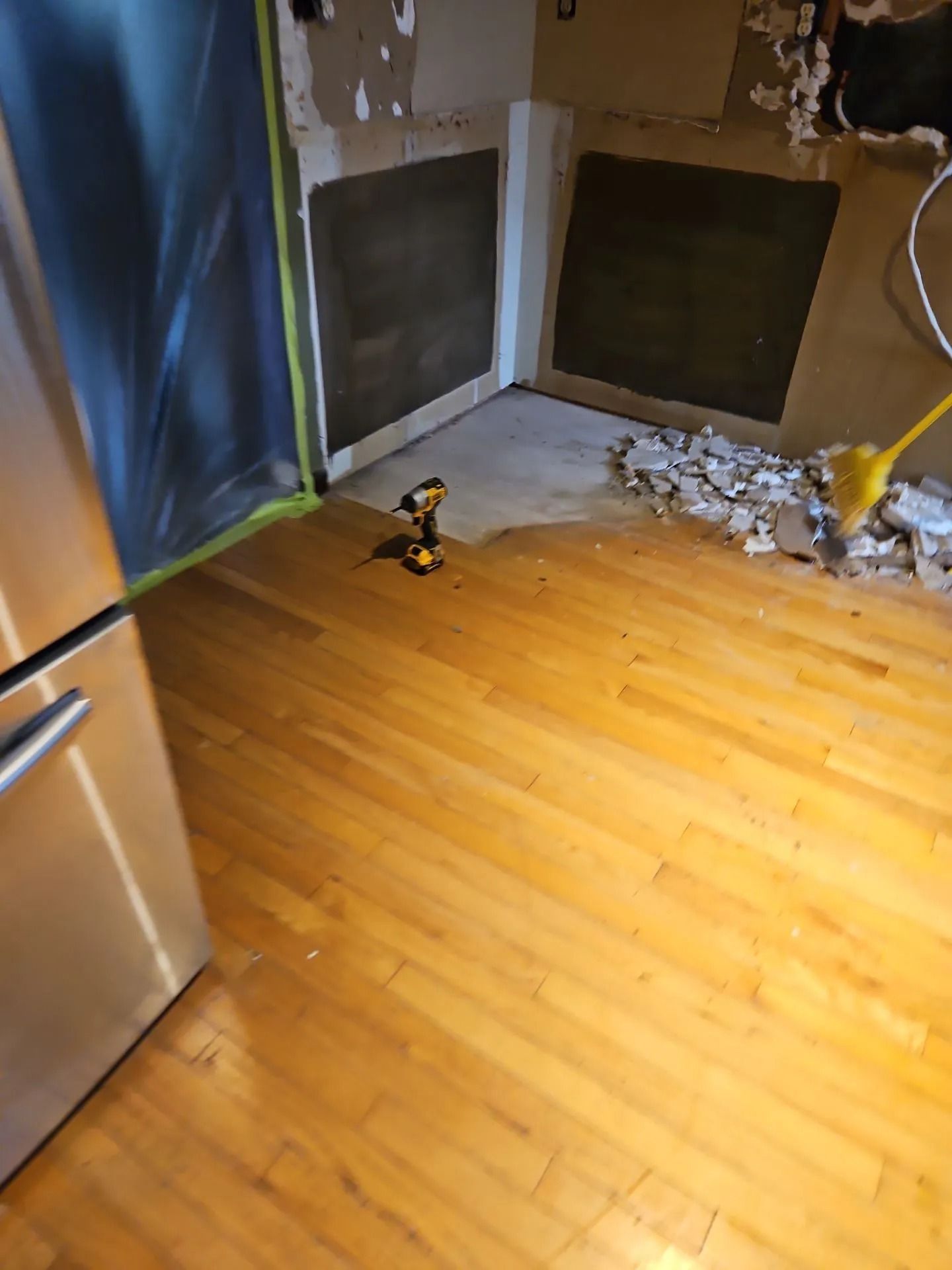 Kitchen undergoing renovation; exposed wall, partially torn, hardwood floor, drill, refrigerator.