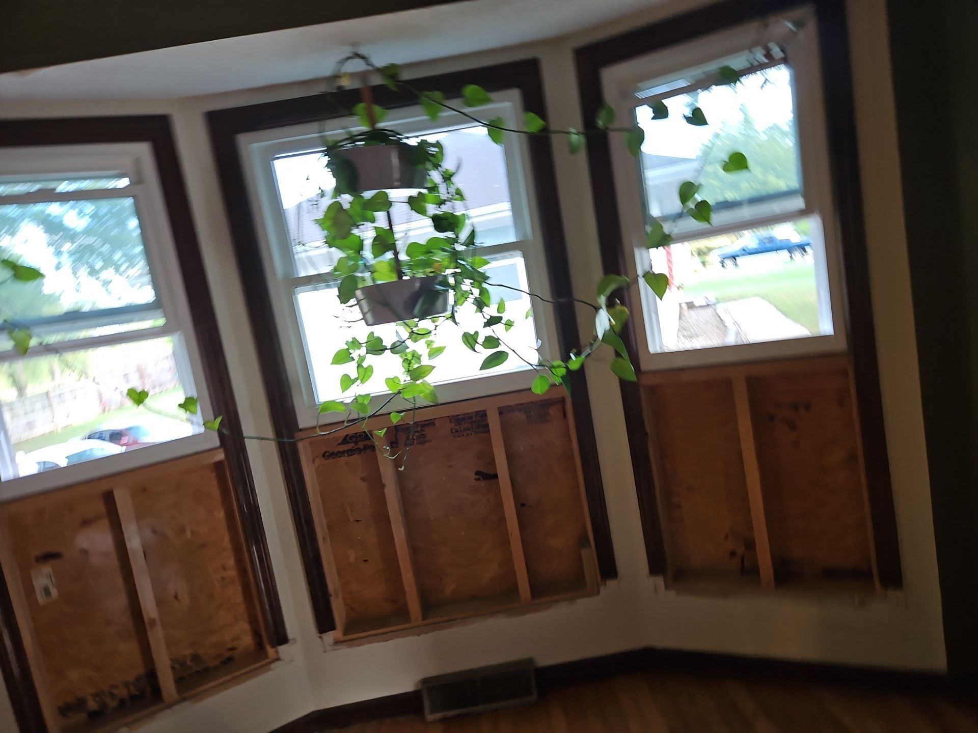 Bay window with three panes, brown trim, exposed wooden framing, and a hanging plant.