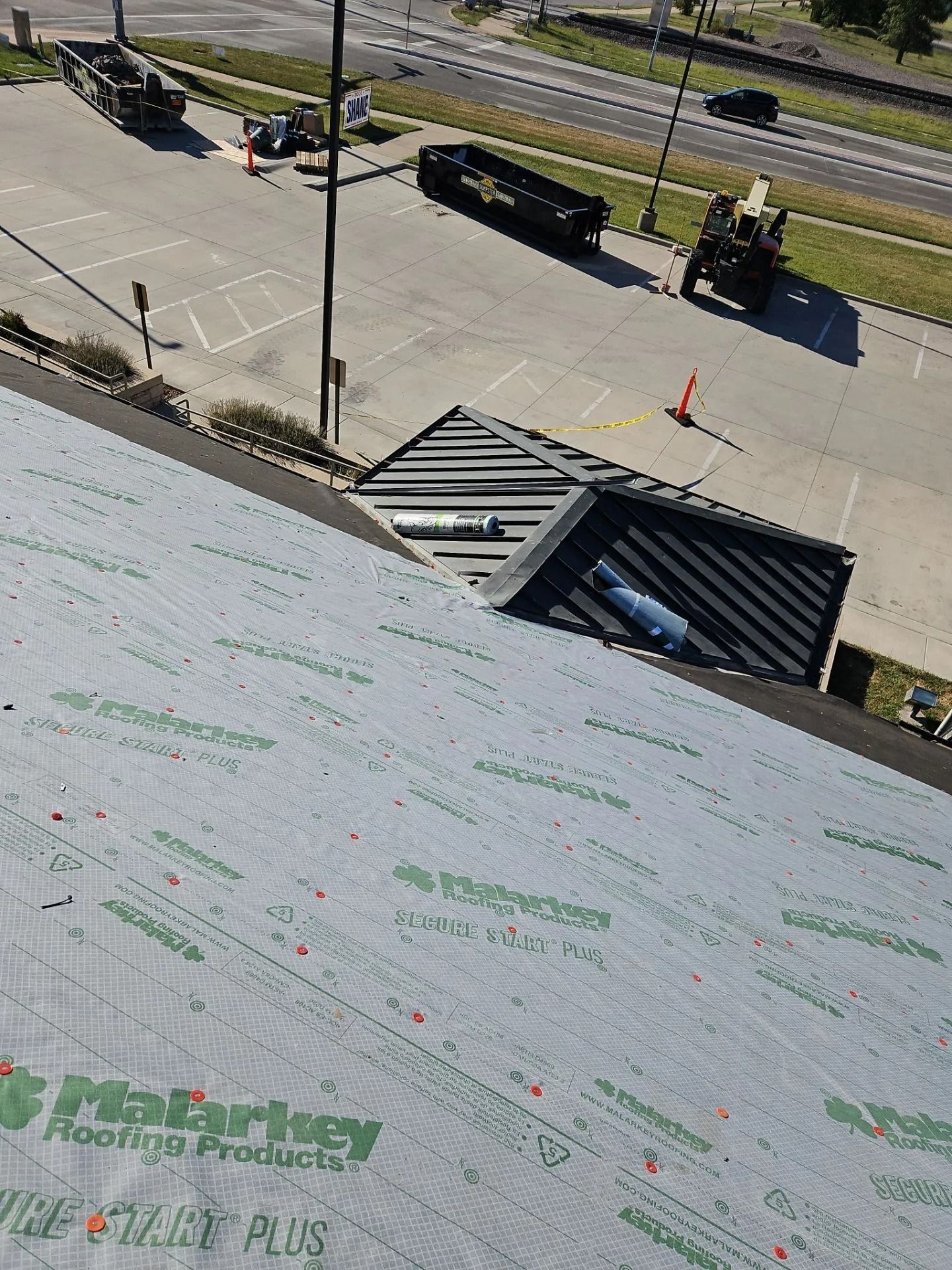 Roofer working on a roof; gray underlayment, black roof, road, equipment, and a parking lot are visible.