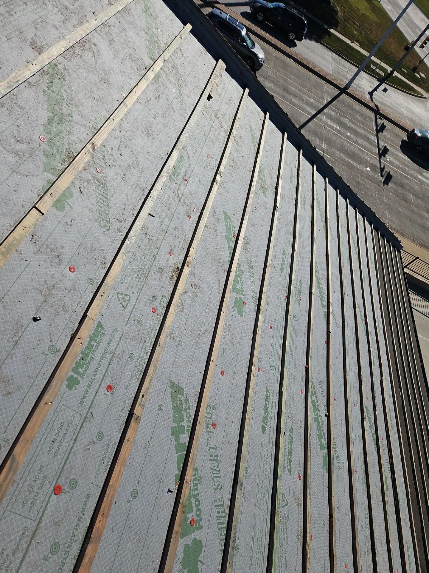View from above of a roof under construction; wooden beams and gray sheathing visible. Street and cars in the background.