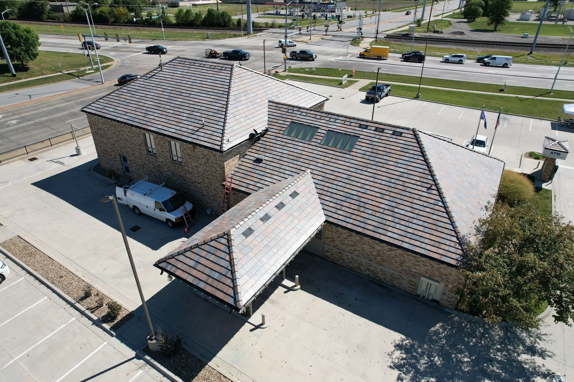 Aerial view of a brown brick building with a light-colored, shingled roof and surrounding parking lot, next to a street with cars.