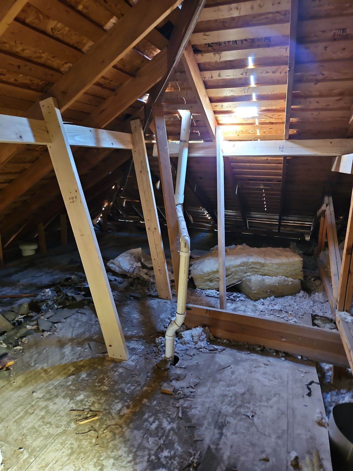 Attic interior with exposed rafters and insulation. White plumbing pipe runs down from roof. Debris on floor.