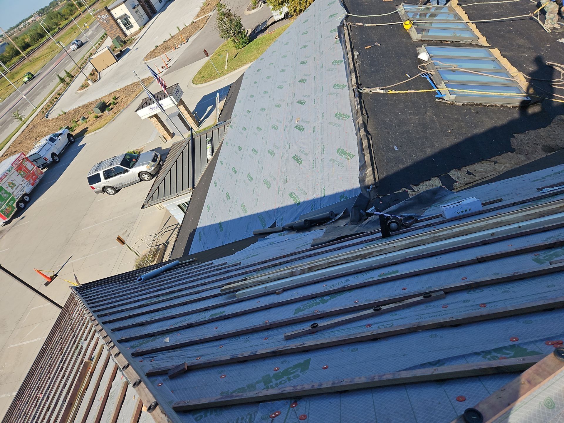Rooftop with metal roofing in progress, view includes vehicles, workers, and buildings.