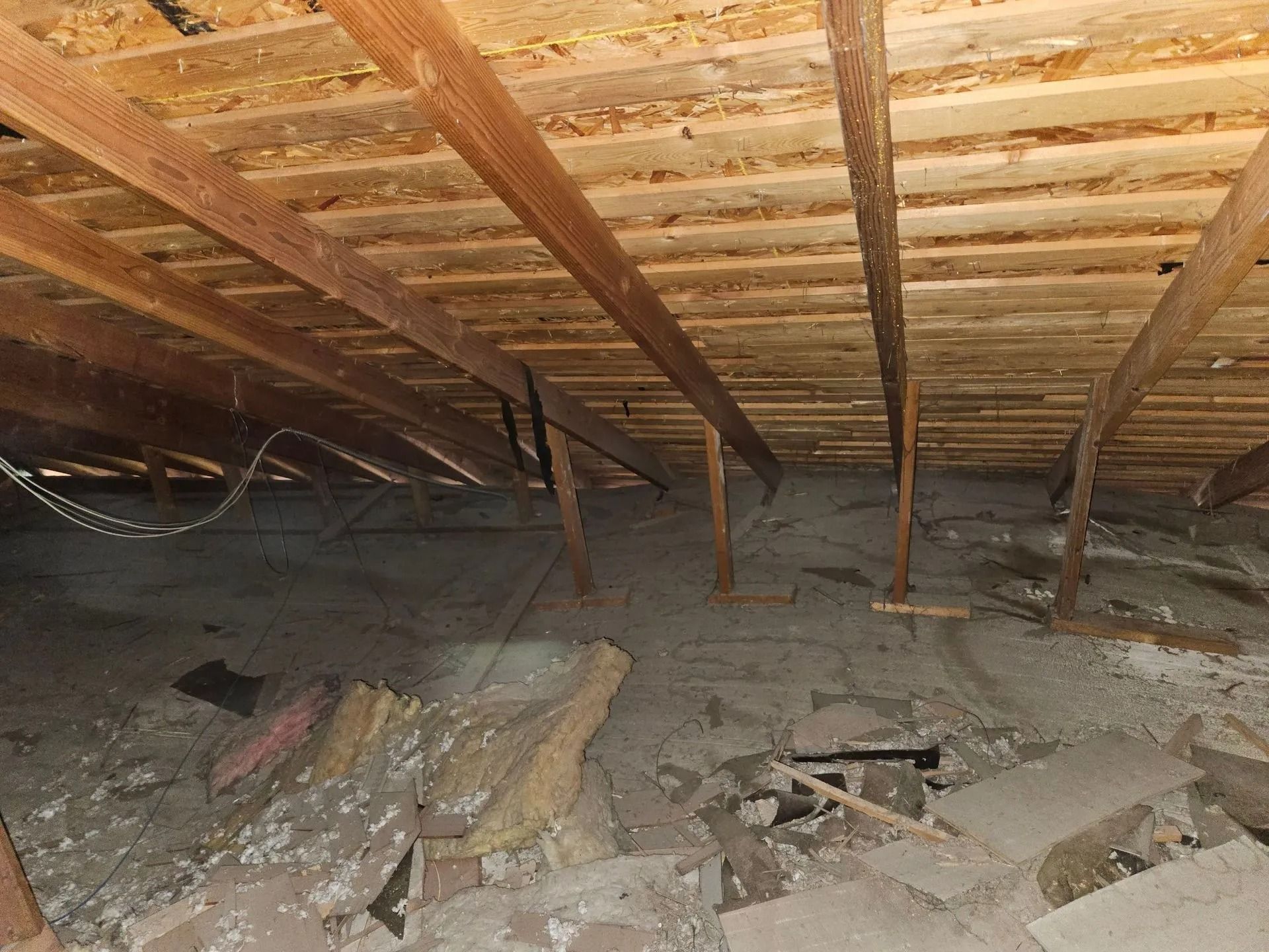 View inside an attic: exposed wooden beams and insulation, debris on the floor.