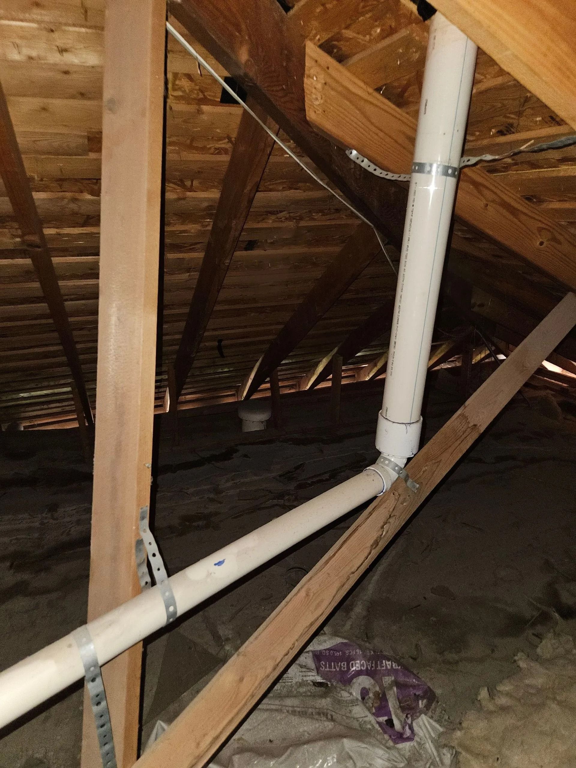 White PVC pipes in attic rafters, held by metal straps. Wooden beams and insulation visible.