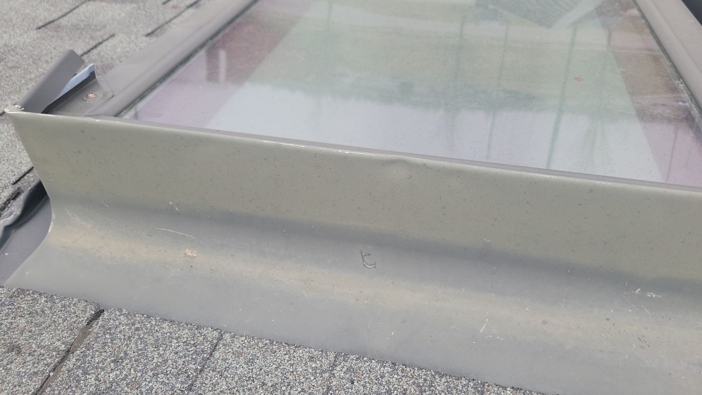 Skylight on a roof; gray metal flashing around the glass, asphalt shingles visible.
