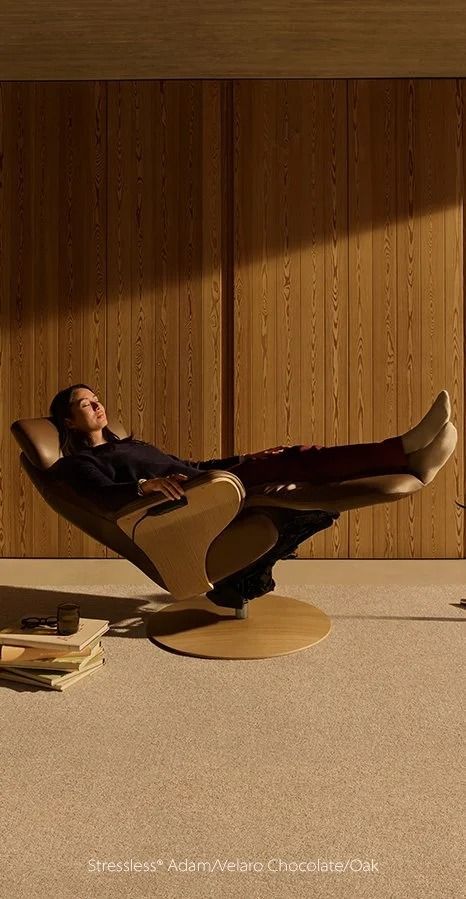 Person reclining in a modern chair in a wood-paneled room, with books stacked on the floor nearby.