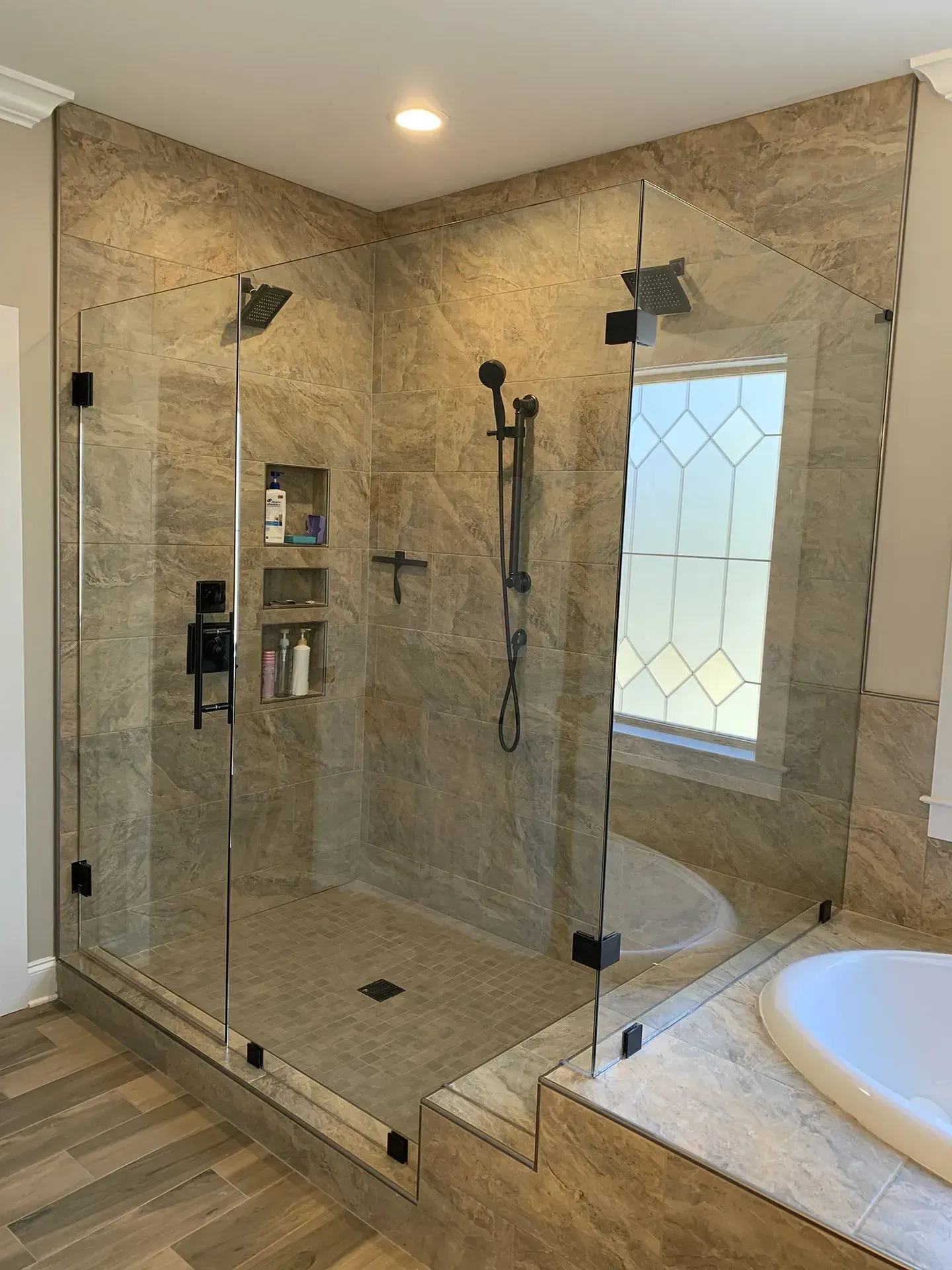 A modern bathroom with a glass-enclosed shower featuring a tan-tiled interior, black fixtures, and a window with a diamond pattern.