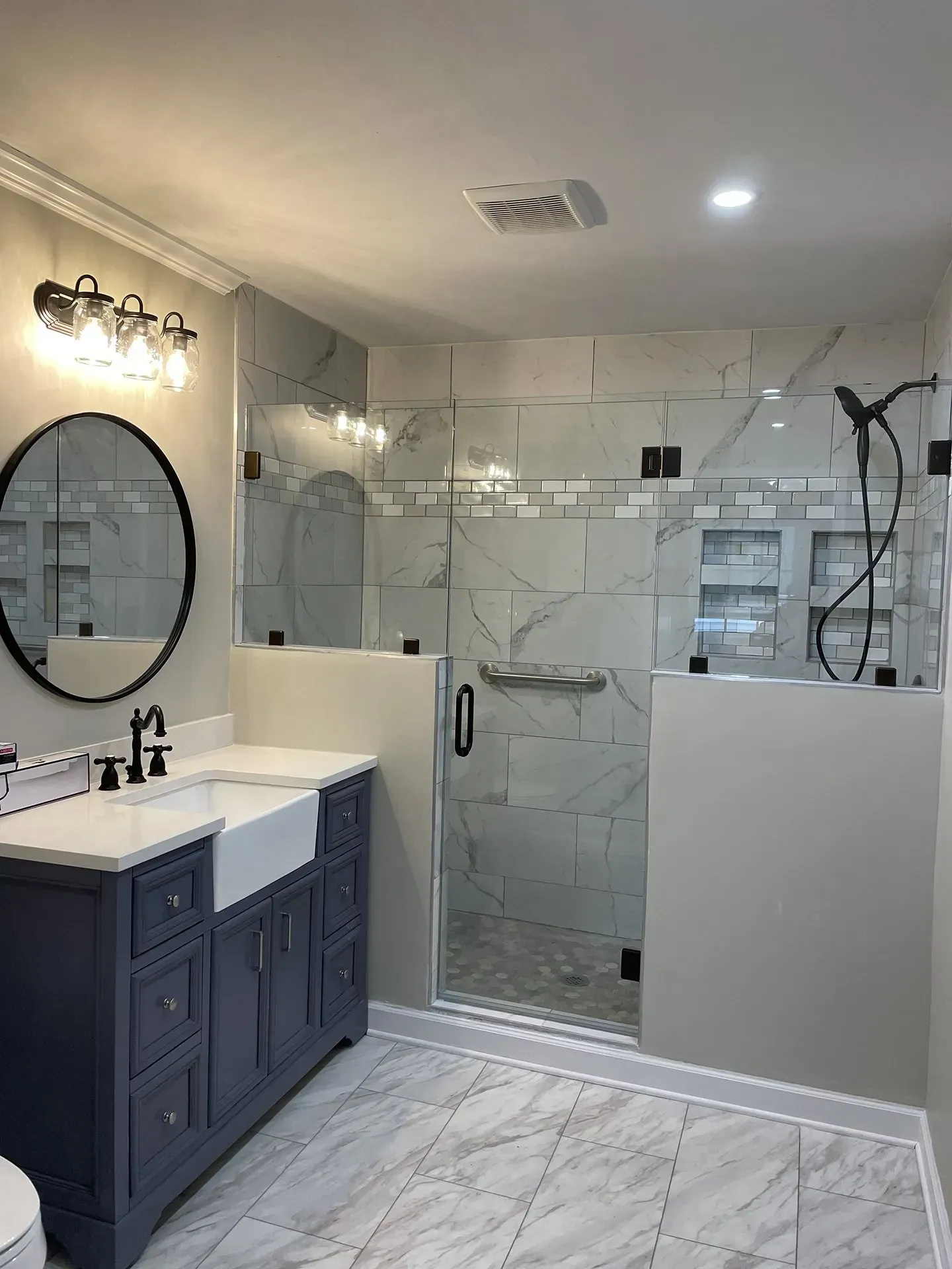 Bathroom with blue vanity, marble-look tile, and a glass-enclosed shower. Features black fixtures and a round mirror.