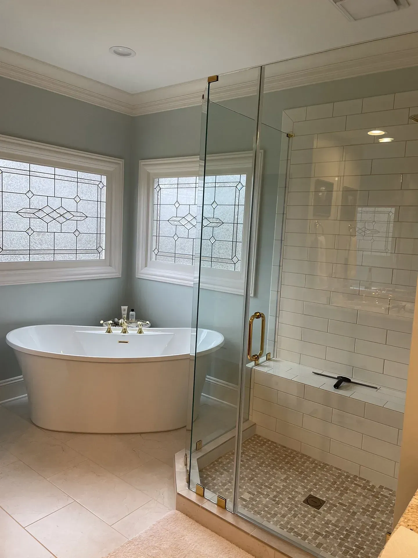 A modern bathroom featuring a white soaking tub, glass shower, and two windows with decorative glass set in a light blue wall.