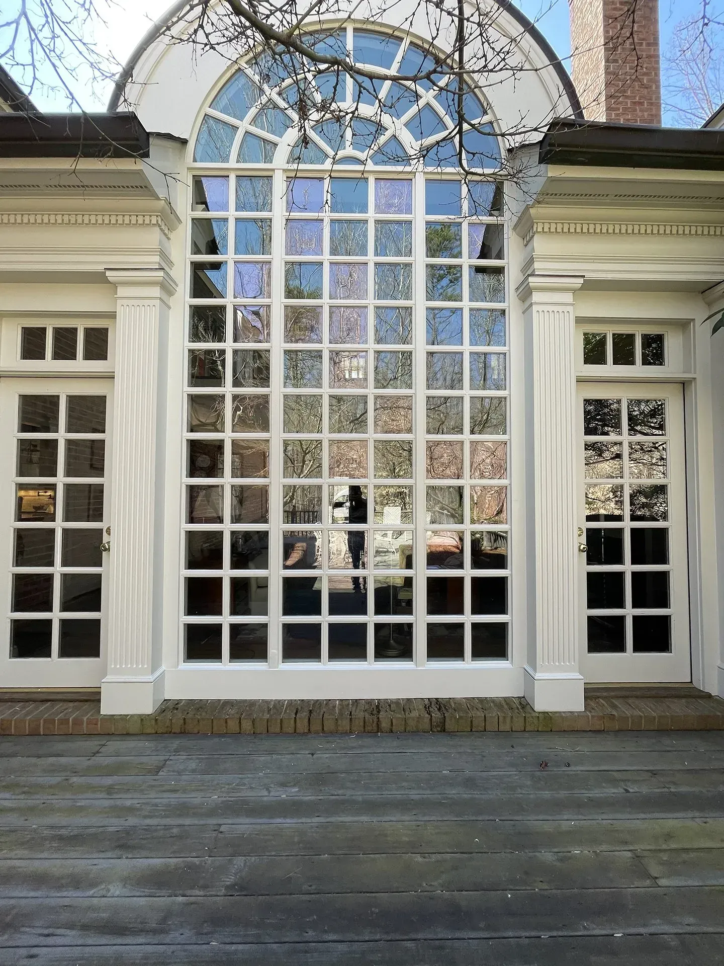 White-framed multi-pane window with arched top and doors on either side, reflecting a room's interior. Set on a stone patio.