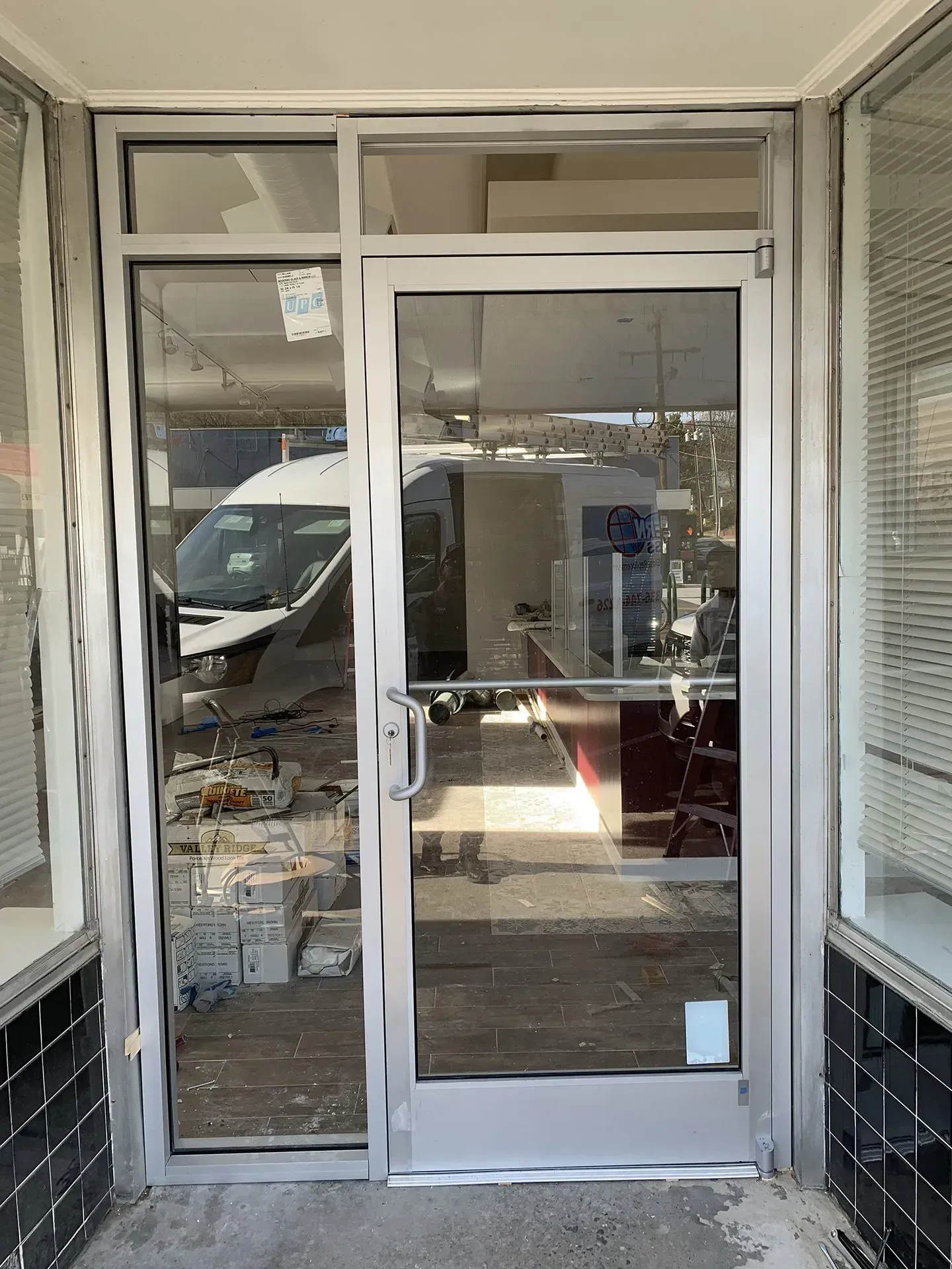 A glass door and sidelight in a silver frame, leading into a partially empty interior. The building's exterior has a tiled facade.