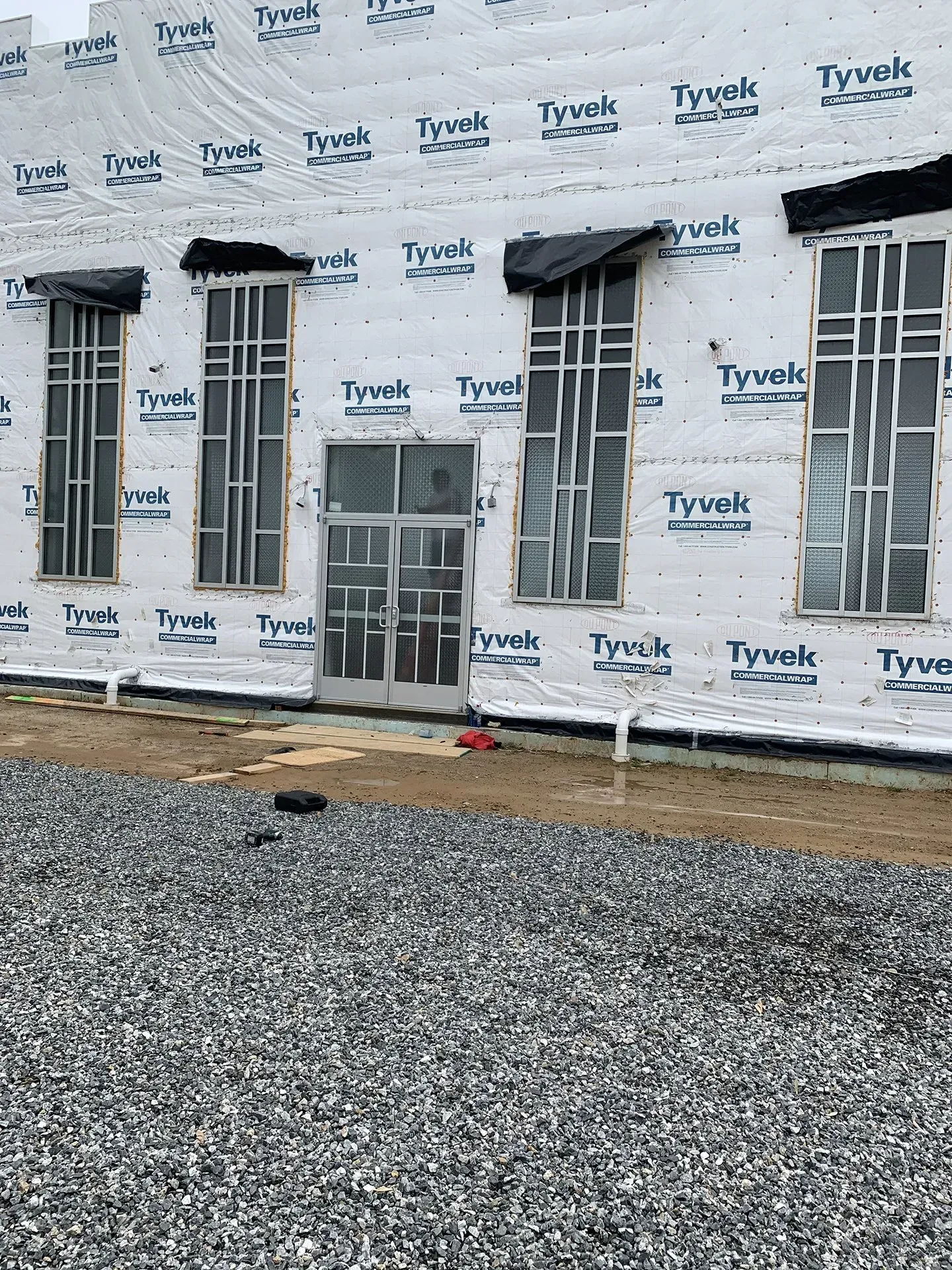 Exterior of a building under construction. Windows and a door are framed and covered with Tyvek.  Gray gravel covers the ground.