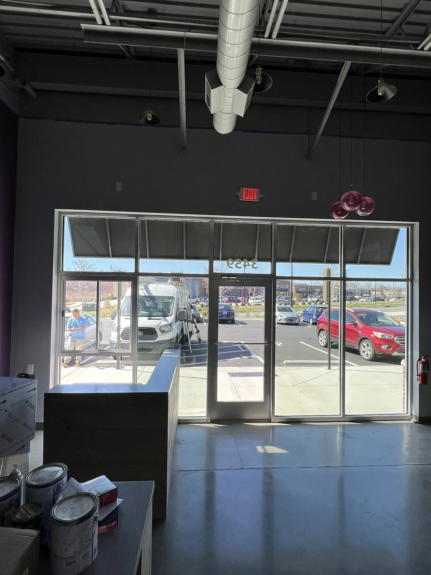 View from inside a commercial space, looking out through large windows. Outside, there are cars and a person. Gray walls and a silver duct are visible.