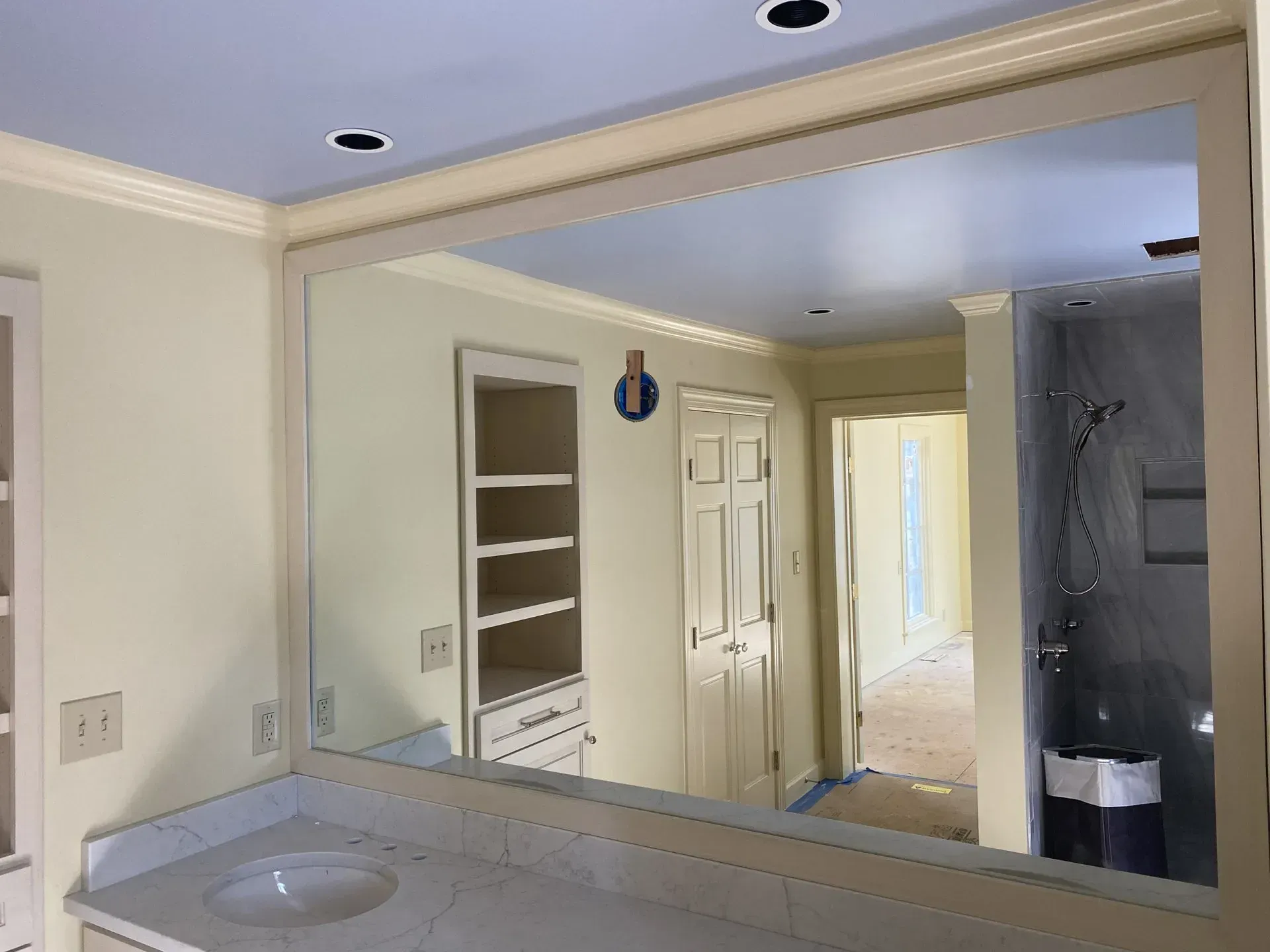 Bathroom with a large rectangular mirror over a white countertop with a sink. A built-in cabinet and a doorway are reflected in the mirror.