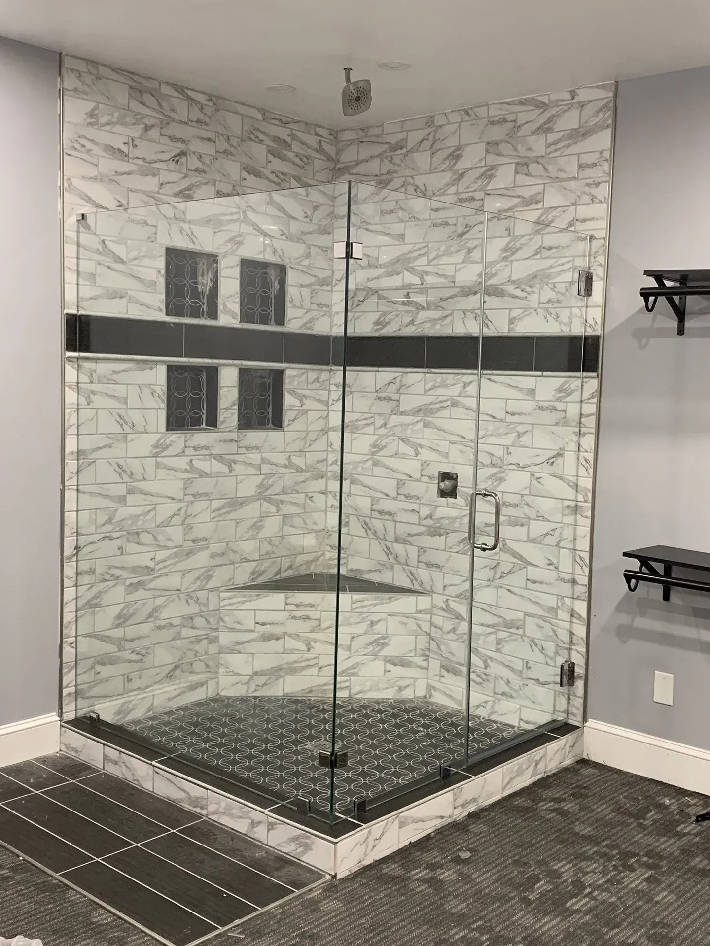 A modern, corner shower with marble-like tile, a black mosaic floor, and glass doors. Grey walls are in the background.