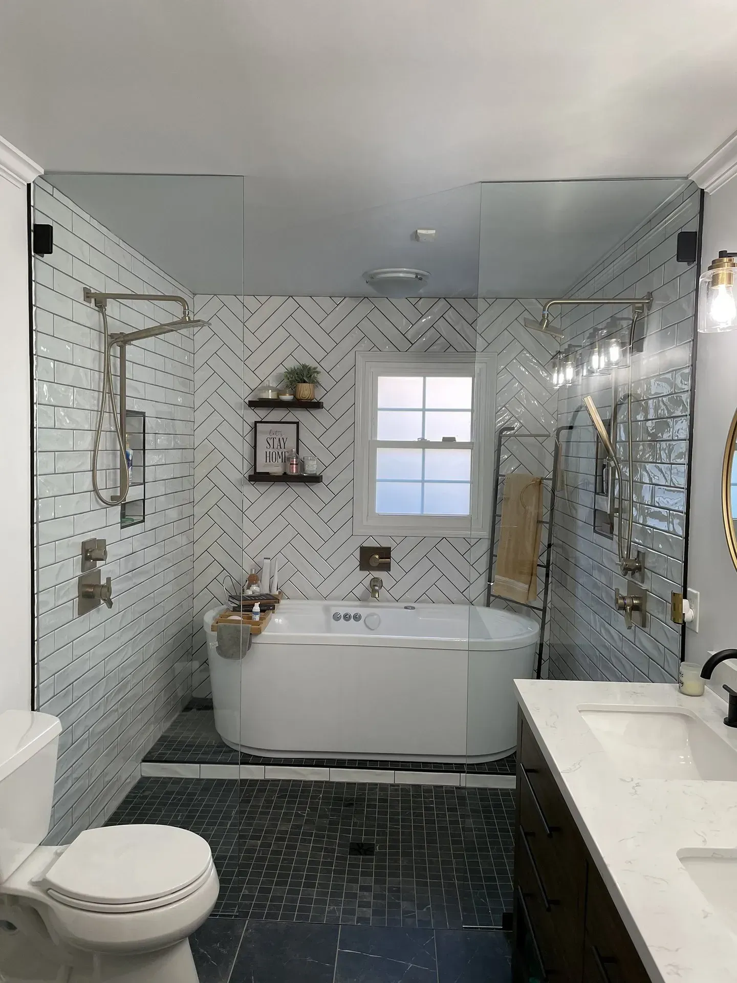 Modern bathroom with a white soaking tub, glass shower enclosure, and herringbone tile wall. Dark floor, white vanity, and a window.