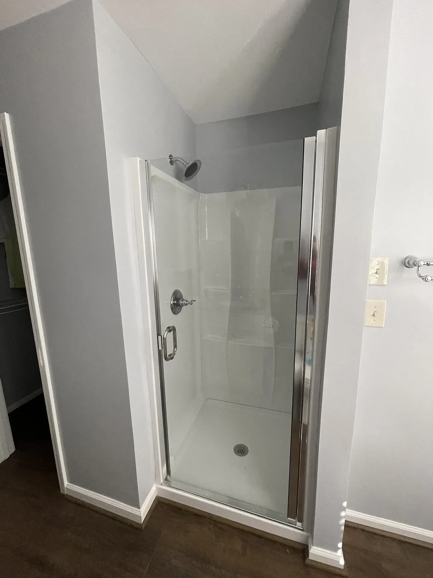 A small, white shower with a glass door is set in a bathroom with gray walls and brown flooring.