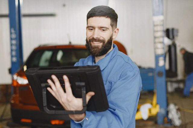 Mechanic in blue shirt inspecting a tablet in an auto repair shop, with a car in the background