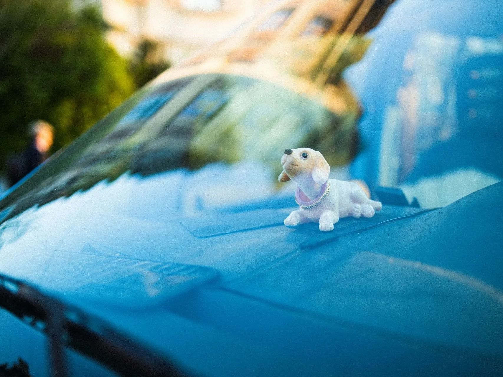Small white dog figurine on a blue car hood, with a blurred outdoor reflection in the windshield