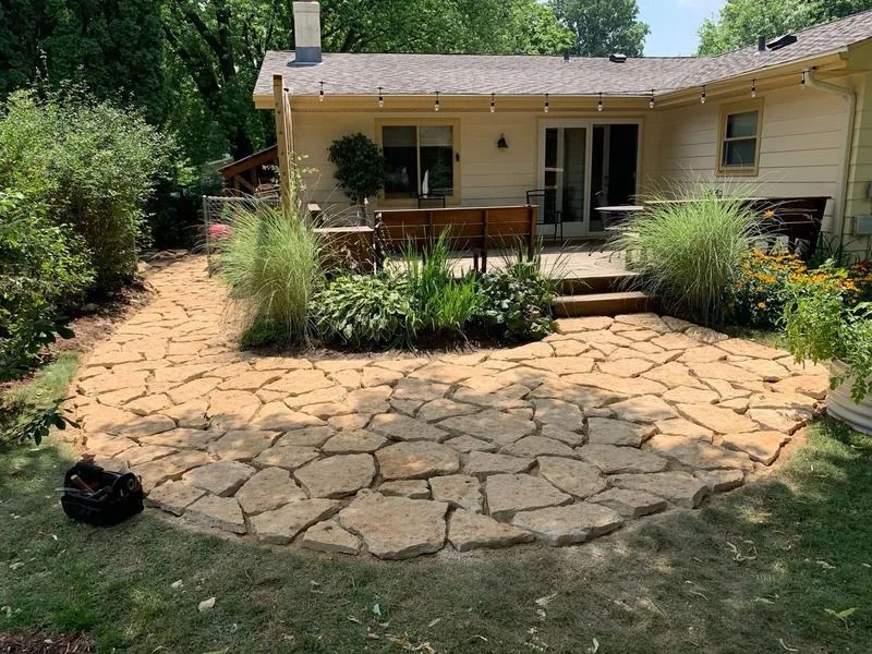 Stone patio with potted plants and tall grasses in a backyard, leading to a beige house with a porch.