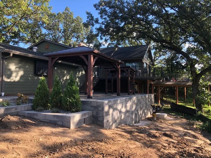 Wooden pergola on a raised stone patio beside a house, with trees and a dirt yard in daylight
