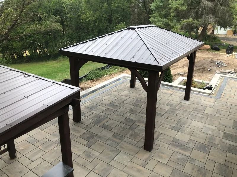 Covered outdoor patio with dark metal roofs and stone pavers beside a grassy, wooded area