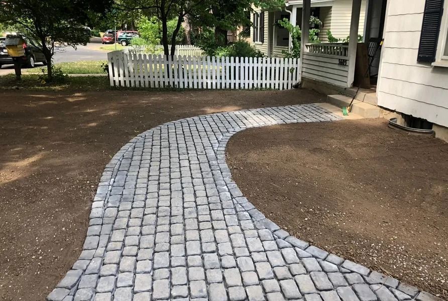 Curved gray brick walkway beside a house, leading to a white picket gate and porch.