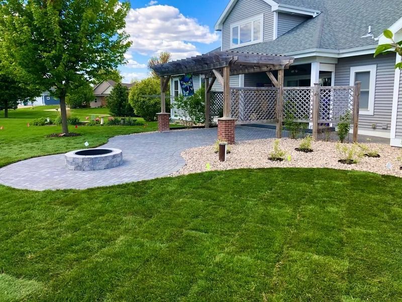 Backyard patio with fire pit, pergola, and curved stone path beside a house and green lawn