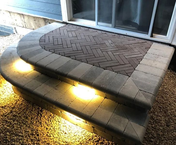 Curved stone front steps and brick patio beside a sliding glass door, lit by two warm ground lights.