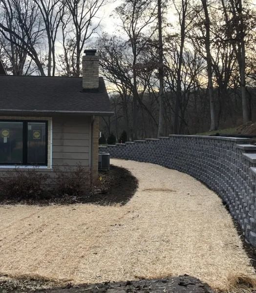 Curved gravel path beside a house, bordered by a stone retaining wall and leafless trees