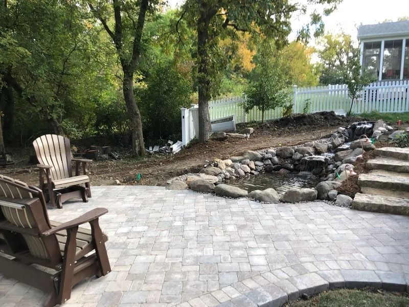 Patio with Adirondack chairs beside a landscaped stone stream and white fence under trees