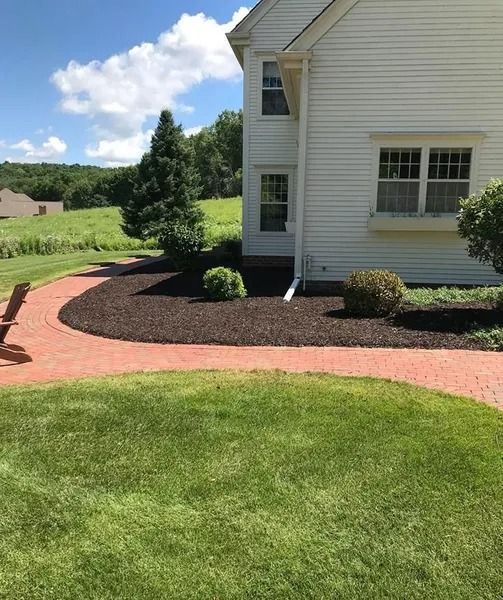 Brick walkway curves through a green lawn beside a white house with dark mulch landscaping.