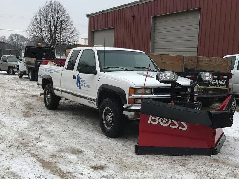 White pickup with front snowplow parked on snowy lot by a red building and another truck behind it