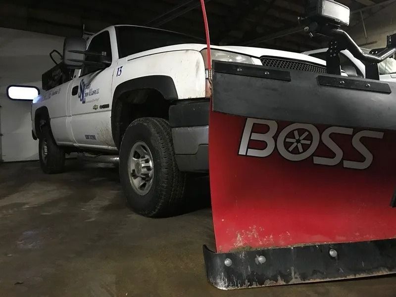 White pickup truck with a red BOSS snow plow in a garage, viewed from the front-left side