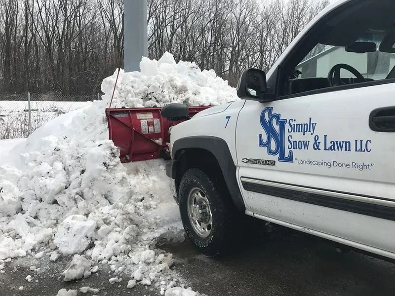 Snow plow attached to a white landscaping truck pushing snow in a winter roadside scene.