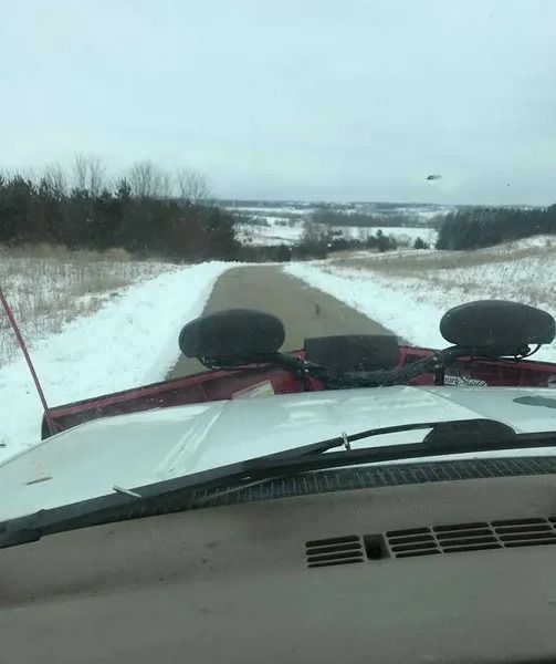 View from a vehicle driving down a snowy rural road with fields and trees ahead.