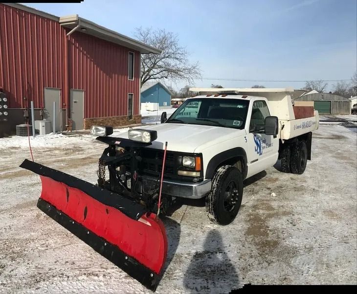 White pickup with red snowplow parked in a snowy yard beside a red barn