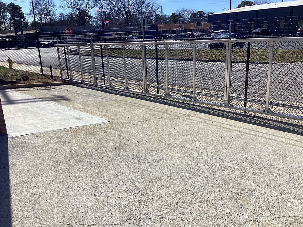 Concrete walkway alongside chain-link fence with metal posts. Street and cars visible in the background.