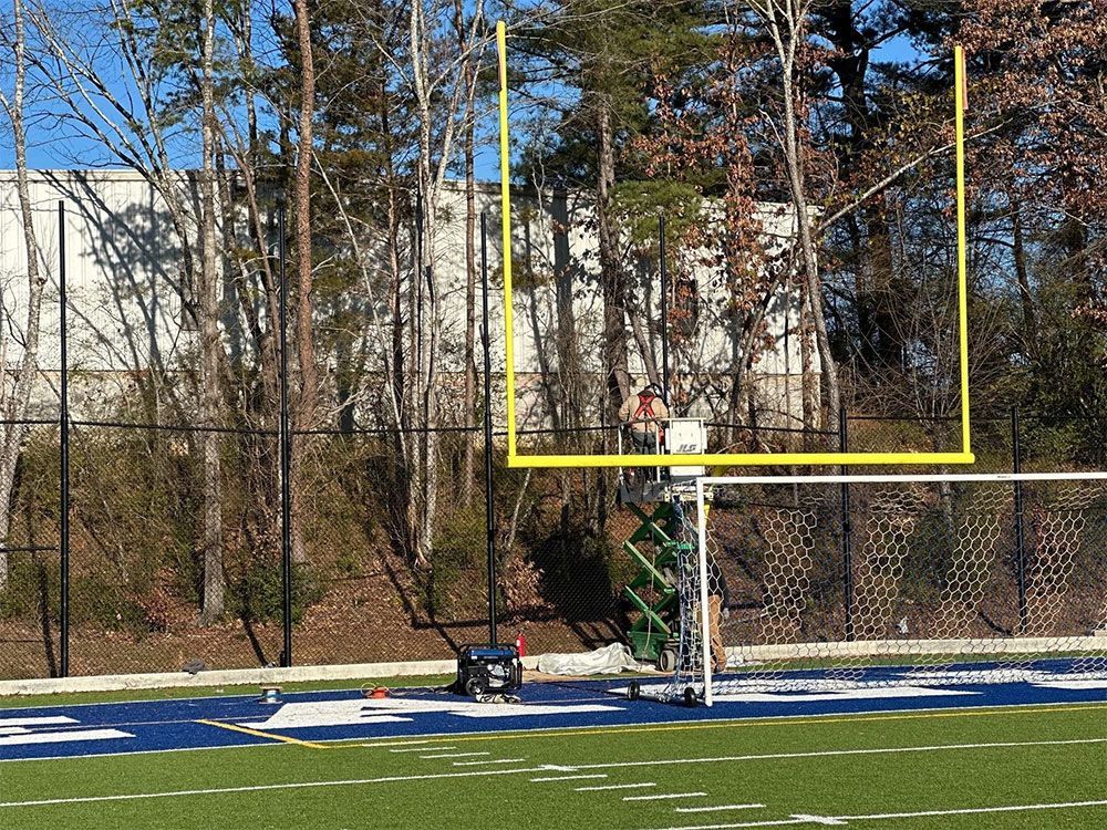 Football field with yellow goalpost. Green turf with white yard lines. Trees and a building in the background.