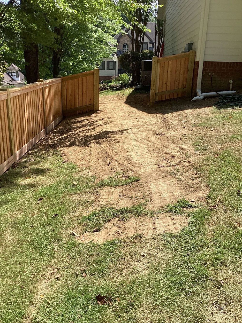 A cleared dirt path leading through a wooden fence gate in a backyard with green grass.