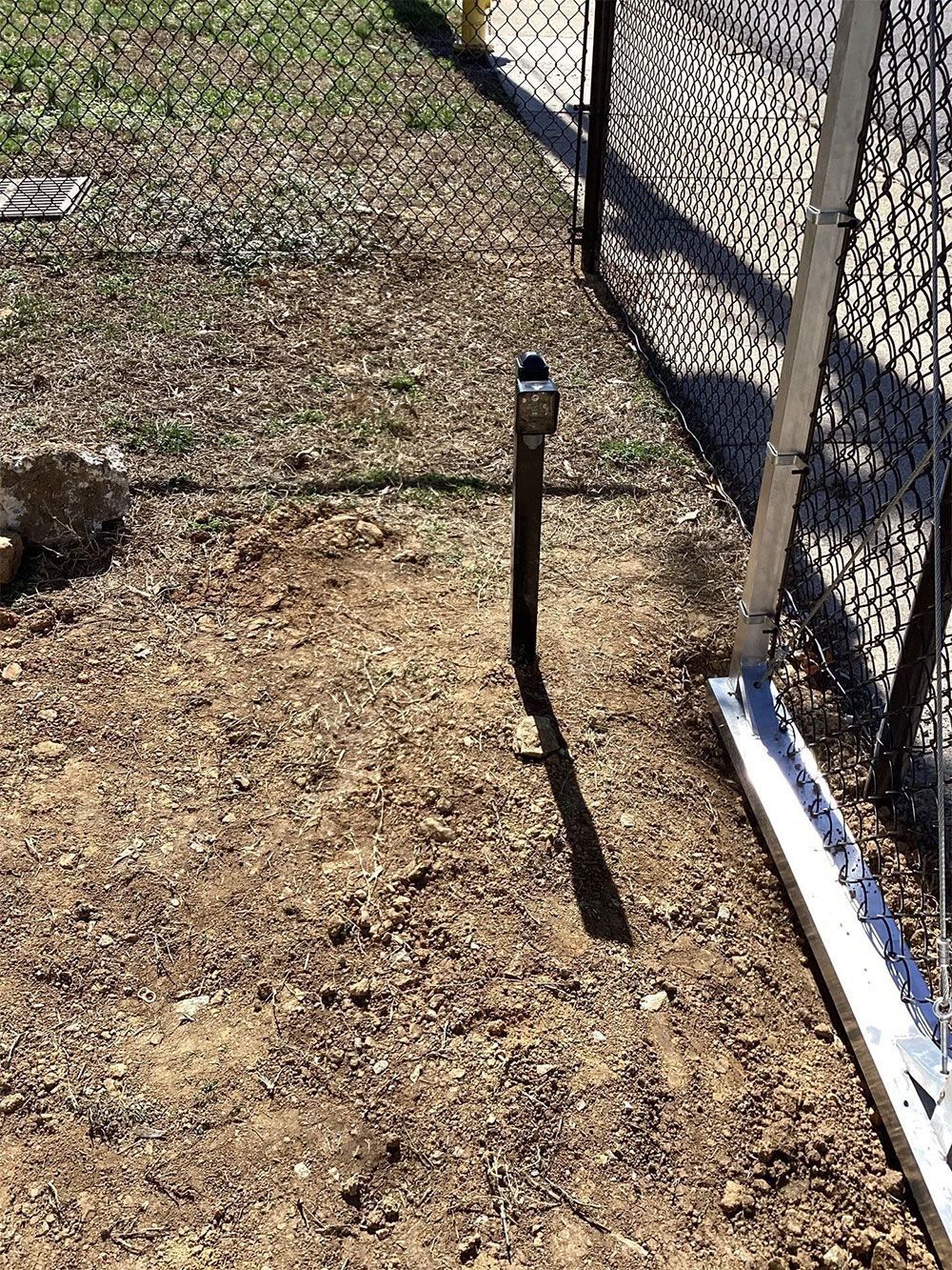 A metal pipe with a cap stands next to a chain-link fence on a dirt ground.