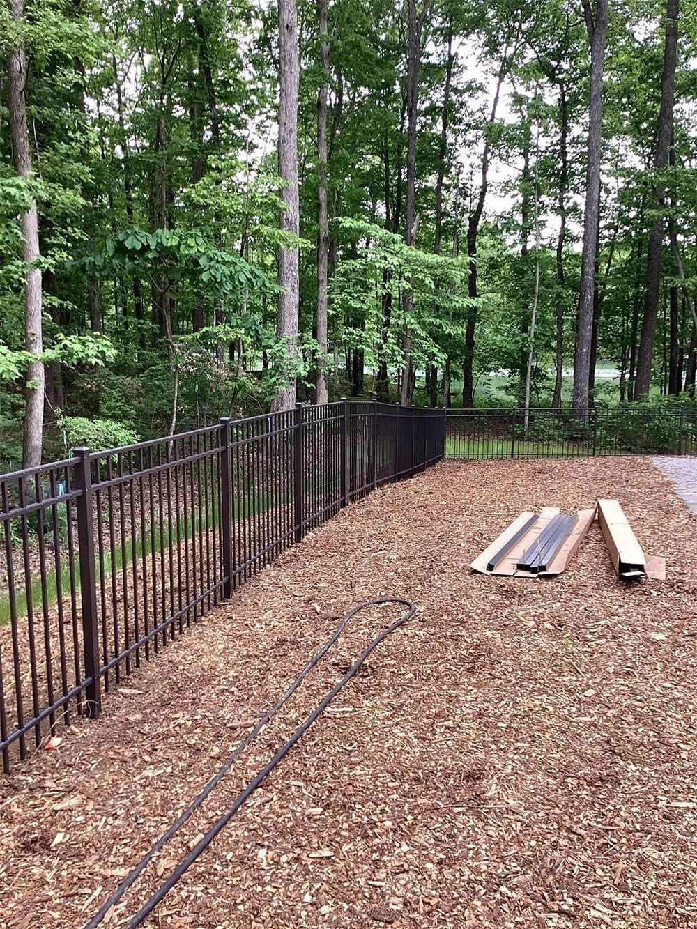 A brown metal fence borders a wood-chipped area next to a forest.