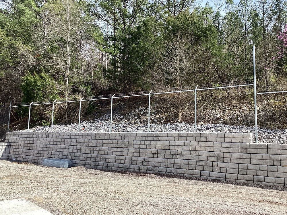 Retaining wall made of gray blocks with a chain-link fence on top, in front of trees.