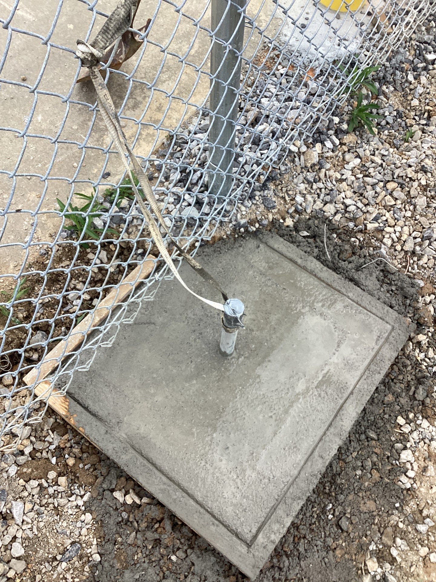 Concrete square base around a metal pole next to a chain link fence.
